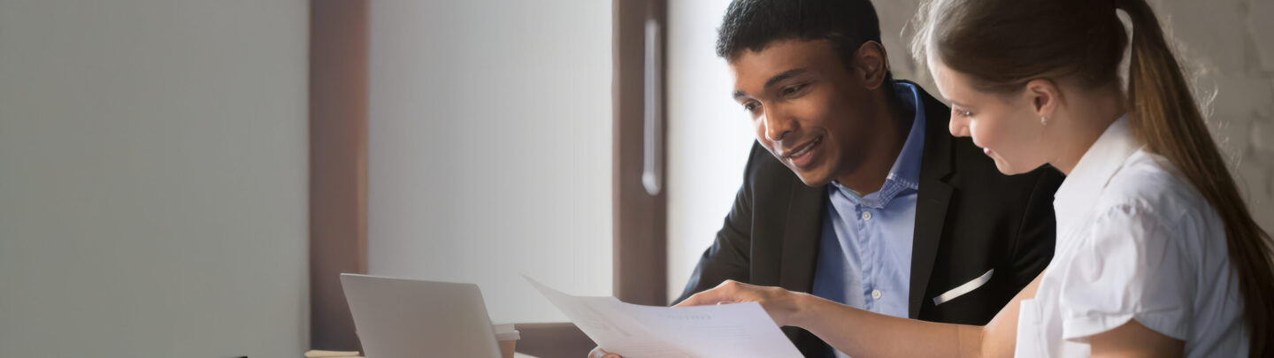 Two people sit at a desk, looking at documents together. One person points at the papers while the other listens. A laptop and coffee cup are on the desk in a well-lit office setting.