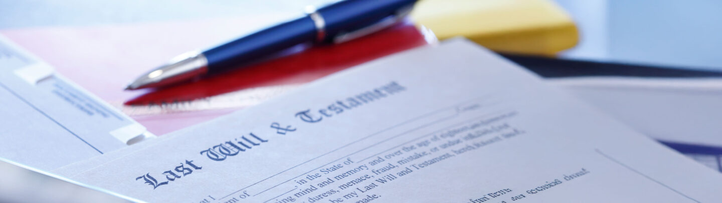 A close-up of a “Last Will & Testament” document on a desk, with a blue pen and some folders in the background. The focus is on the title of the document.