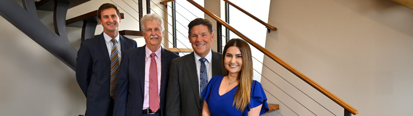 Four professionally dressed people stand smiling in front of a modern staircase with metal railings in a well-lit indoor setting.