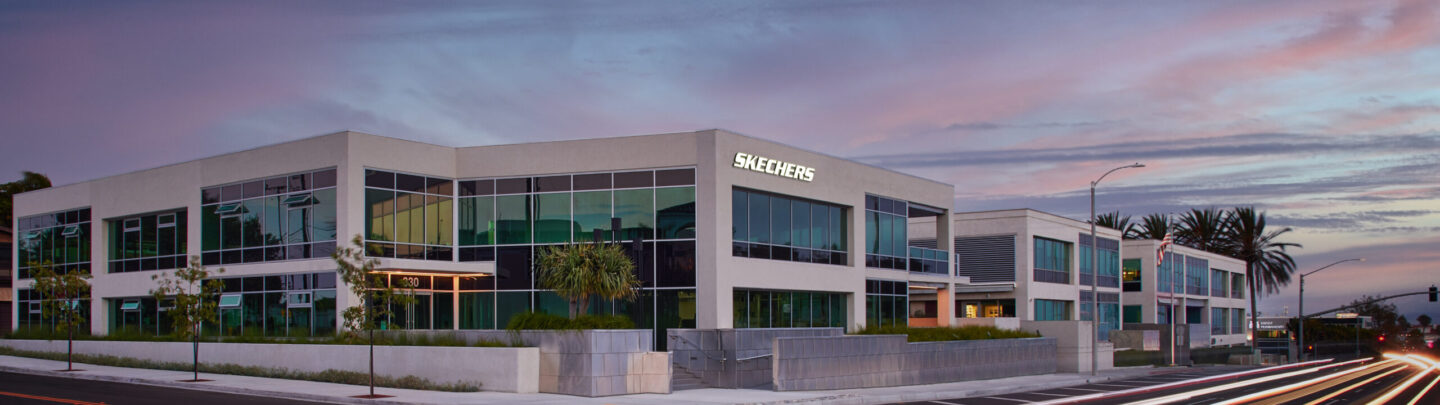 A modern office building with large windows and a sign reading “Skechers” is shown at dusk. Light trails from passing vehicles are visible on the street in the foreground. Palm trees line the property.