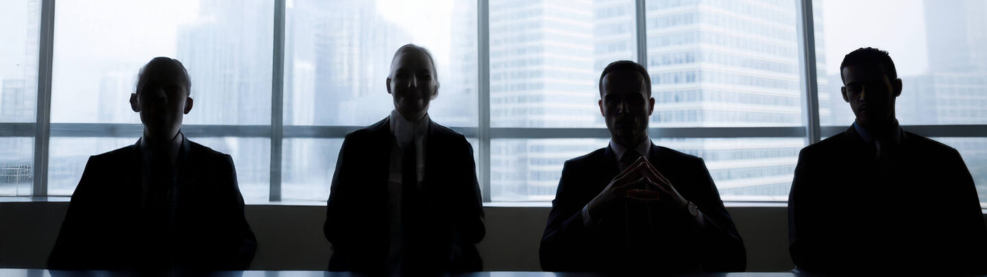 Four people in business attire sit at a table in front of large windows, their faces and bodies silhouetted against the bright cityscape outside.