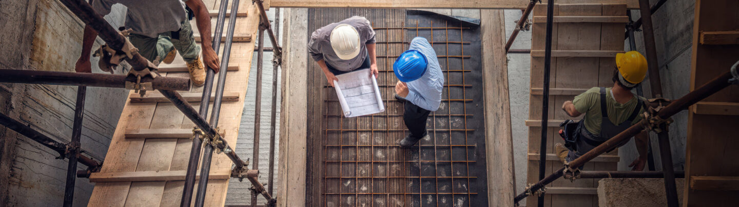 Aerial view of construction workers wearing helmets on scaffolding and stairs, with two people in the center reviewing blueprints on a rebar-lined concrete floor.