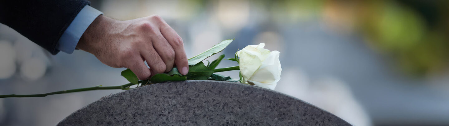 A person’s hand gently places a white rose on a gray tombstone, symbolizing remembrance and mourning in a cemetery with a blurred background.
