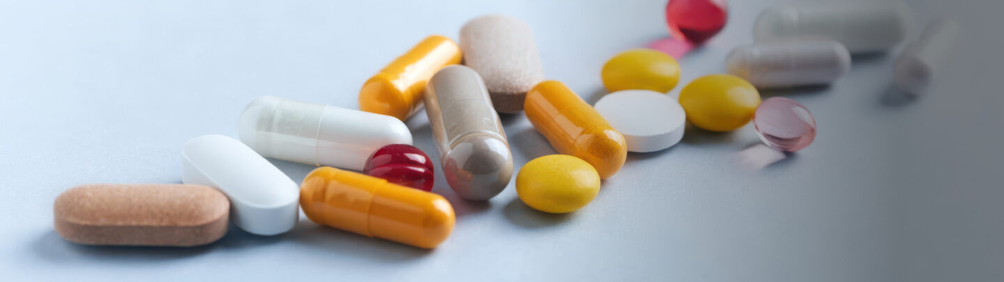 A variety of pills and capsules in different shapes, sizes, and colors scattered on a light surface, including yellow, white, brown, orange, and red tablets.