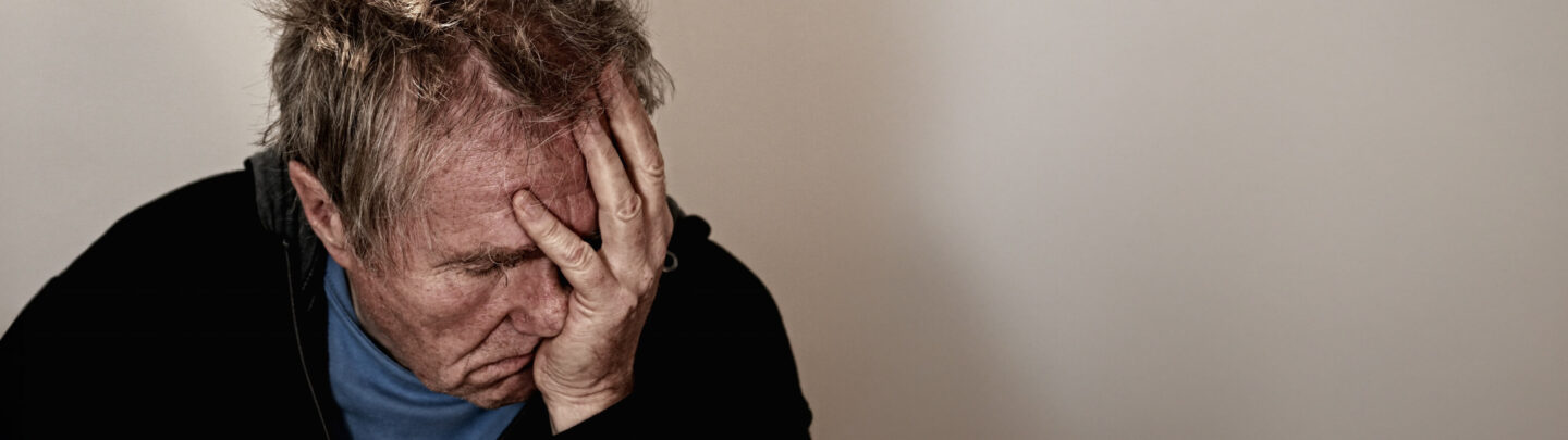 An older man with gray hair sits with his hand covering his face, appearing distressed or overwhelmed, against a plain light background.