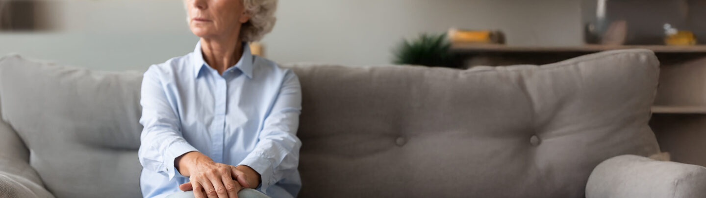 An older woman with gray hair sits on a gray sofa, looking thoughtfully to the side with her hands resting on her knee. The background shows a blurred indoor living space.