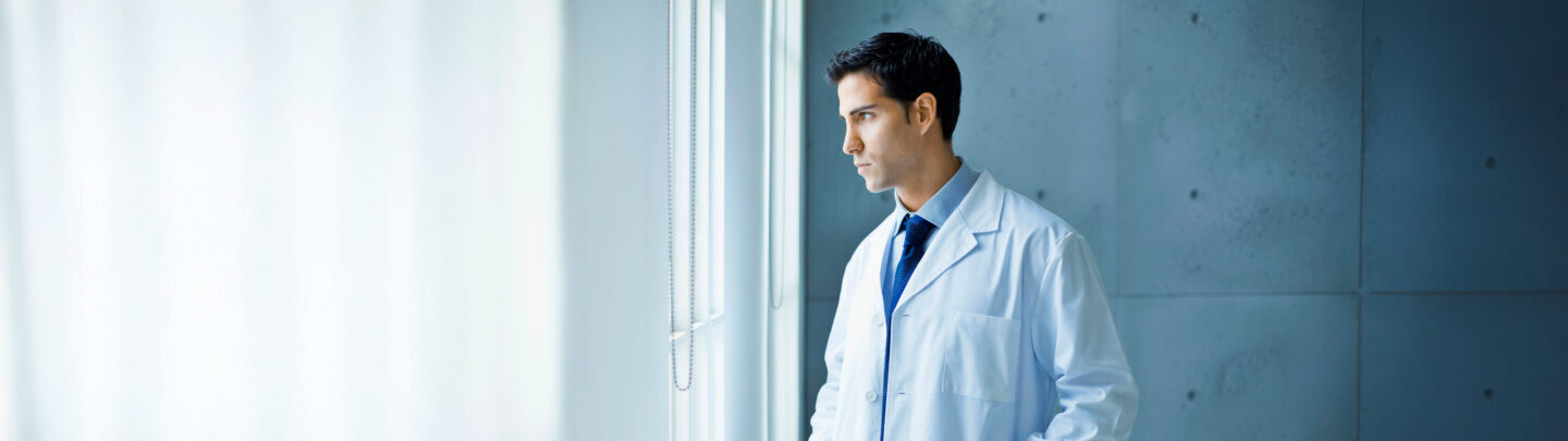A man in a white lab coat and blue tie stands indoors by a window, looking outside with a thoughtful expression. The background is softly lit, with blue and white tones.