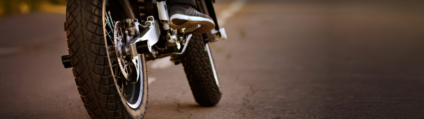 Close-up of a person wearing sneakers riding a motorcycle on a paved road, with focus on the rear wheel and part of the rider’s foot, background blurred.