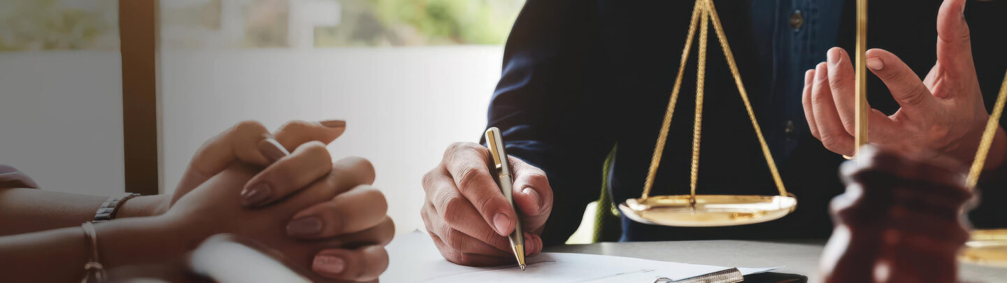 Two people sit at a table with legal documents, one writing with a pen. A golden scale of justice and a judge’s gavel are visible, suggesting a legal or consultation setting.