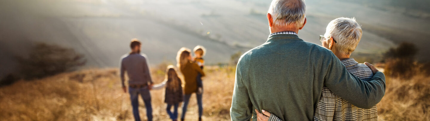 An elderly couple stands on a hill, arm in arm, looking at a family—two adults and two children—walking ahead in a sunlit, rural landscape. The scene is warm and peaceful.
