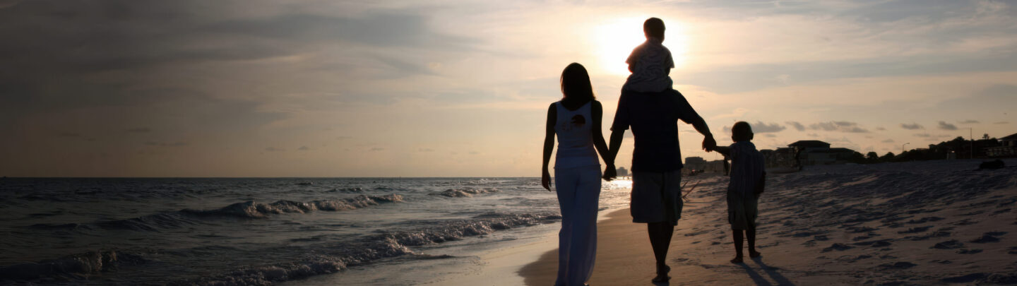 A family of four walks along a sandy beach at sunset. The sun is low in the sky, creating a warm glow and casting silhouettes of the two adults and two children, one riding on an adult’s shoulders. Waves roll in beside them.