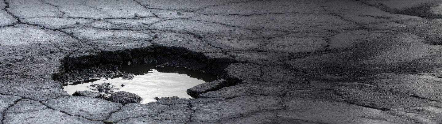 A large pothole filled with water on a cracked, worn asphalt road. The surrounding pavement is visibly eroded and uneven, showing various cracks and rough surfaces.