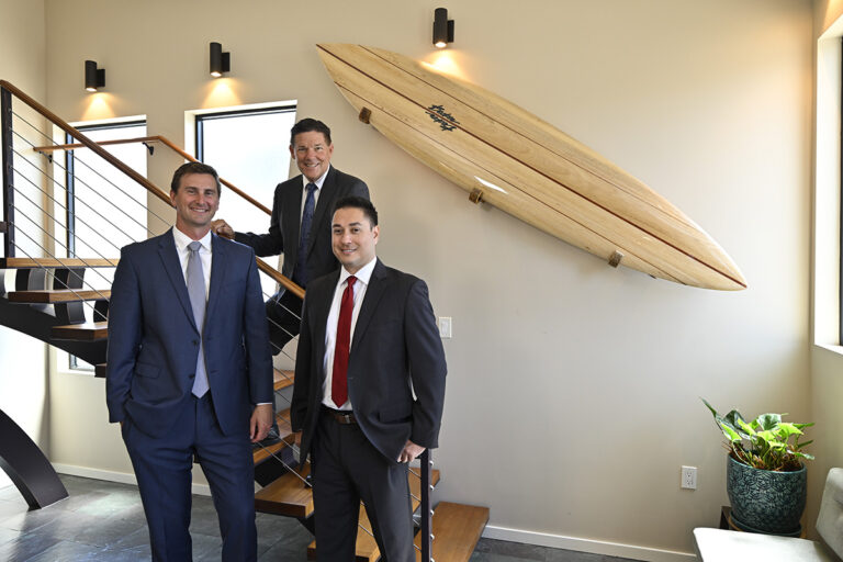 Three men in business suits stand and smile in a modern office space with a staircase and a large wooden surfboard mounted on the wall behind them. There is a potted plant in the corner and sunlight streams through windows.