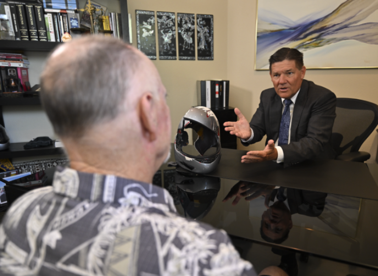 A man in a suit speaks seriously to another man in a patterned shirt across a desk, with a helmet between them, in an office filled with books and framed photos on the wall.