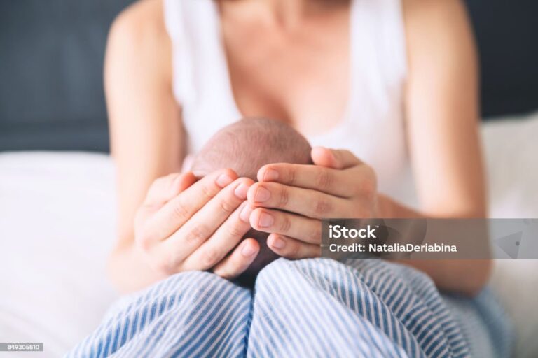 A woman in a white tank top sits on a bed, gently cradling a babys head in her hands, with the baby resting on her lap. Only the back of the babys head and the womans hands are visible.
