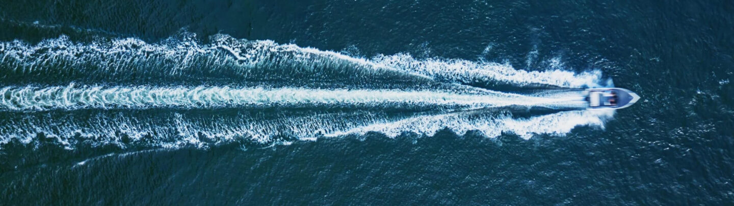 Aerial view of a speedboat moving quickly across deep blue water, leaving a wide, white, V-shaped wake trailing behind it.