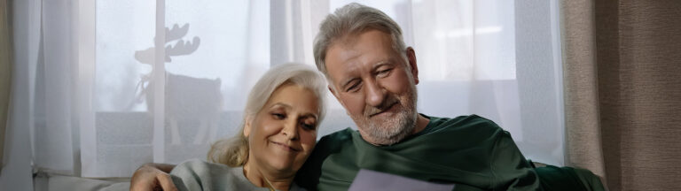 An elderly couple sits closely together on a couch, smiling as they look at a piece of paper, with soft natural light coming through a window behind them.