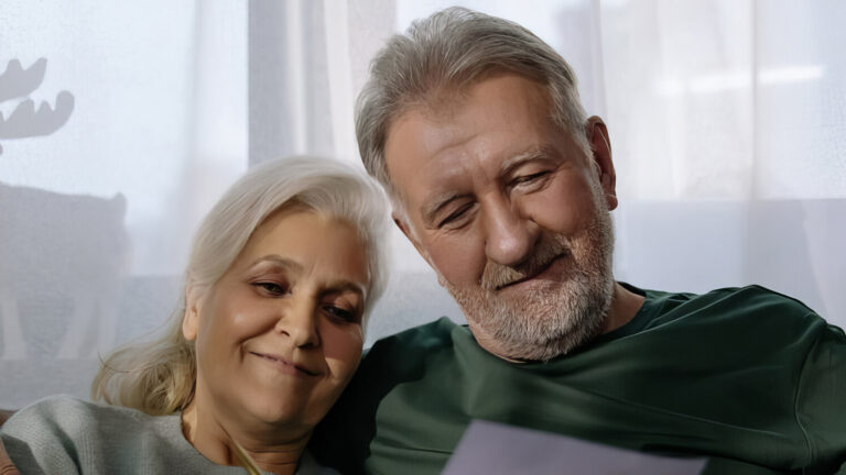 An elderly couple sits closely together on a couch, smiling as they look at a piece of paper, with soft natural light coming through a window behind them.