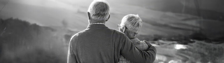 An elderly couple stands closely together outdoors, viewed from behind. The person on the left has their arm around the shoulder of the person on the right. The scene is in black and white with a blurred natural background.