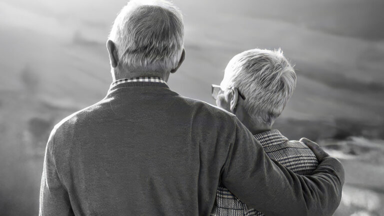 An elderly couple stands closely together outdoors, viewed from behind. The person on the left has their arm around the shoulder of the person on the right. The scene is in black and white with a blurred natural background.