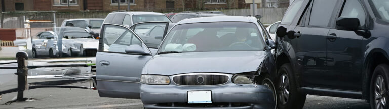 A silver car with a dented front and an open door is involved in a collision with a black SUV on a street; other parked vehicles are visible in the background.