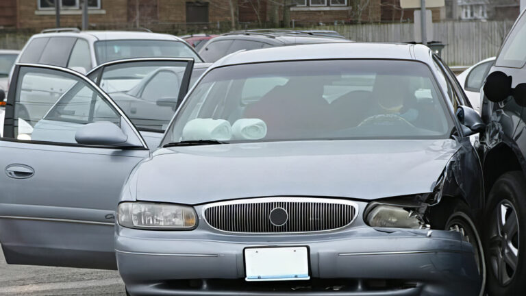 A silver car with a dented front and an open door is involved in a collision with a black SUV on a street; other parked vehicles are visible in the background.