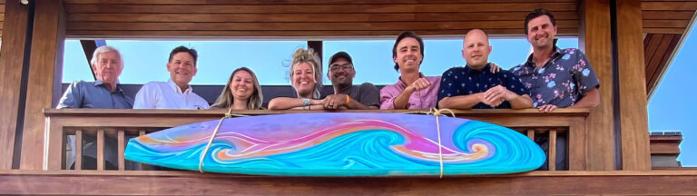 Eight people stand smiling on a wooden balcony behind a colorful surfboard with swirling patterns, under a clear blue sky.