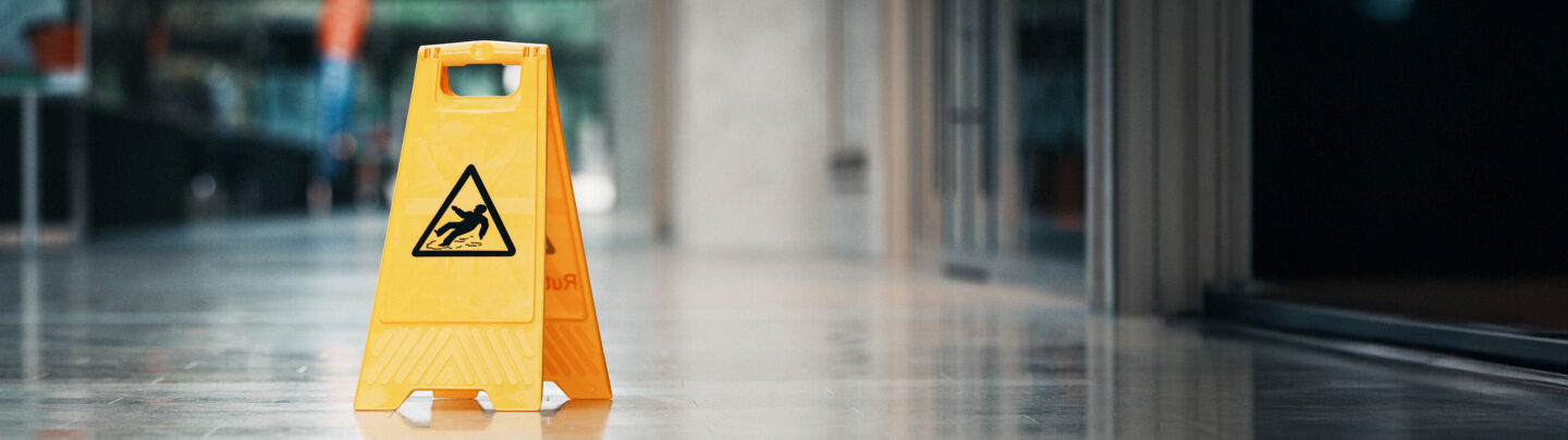 A yellow caution sign with a slipping person icon stands on a shiny indoor floor, warning of a wet or slippery surface. The background shows blurred walls and glass doors in a modern building.