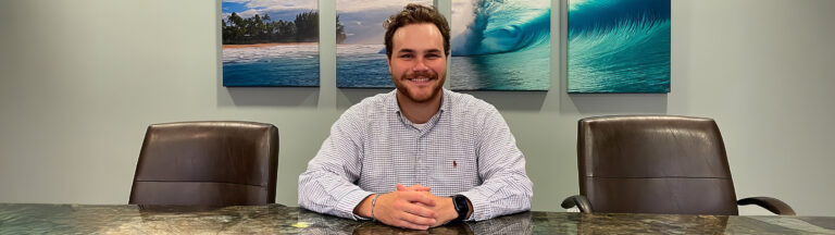 A man with brown hair and a beard sits smiling at a conference table between two empty chairs, with ocean and wave artwork hanging on the wall behind him.
