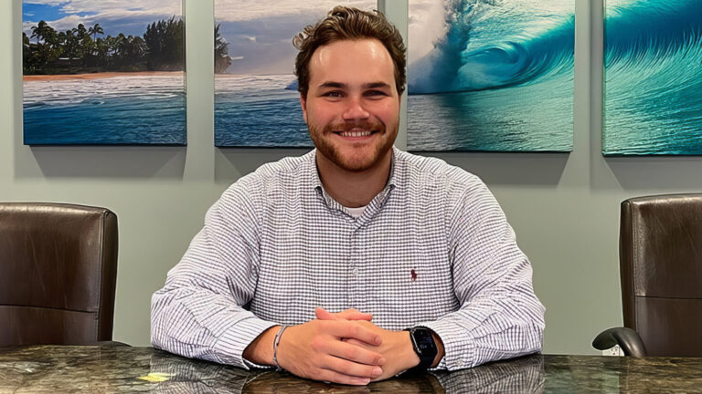 A man with brown hair and a beard sits smiling at a conference table between two empty chairs, with ocean and wave artwork hanging on the wall behind him.