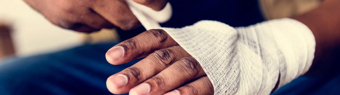 Close-up of a person’s hand being wrapped with a white bandage, suggesting treatment for an injury. The background is blurred, focusing attention on the hand and bandage.
