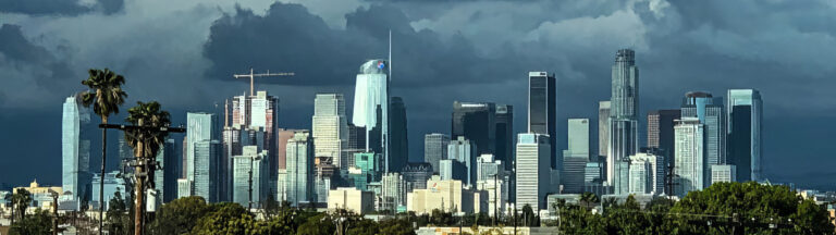 A panoramic view of downtown Los Angeles, featuring tall skyscrapers under a dramatic, cloudy sky, with palm trees and greenery visible in the foreground.