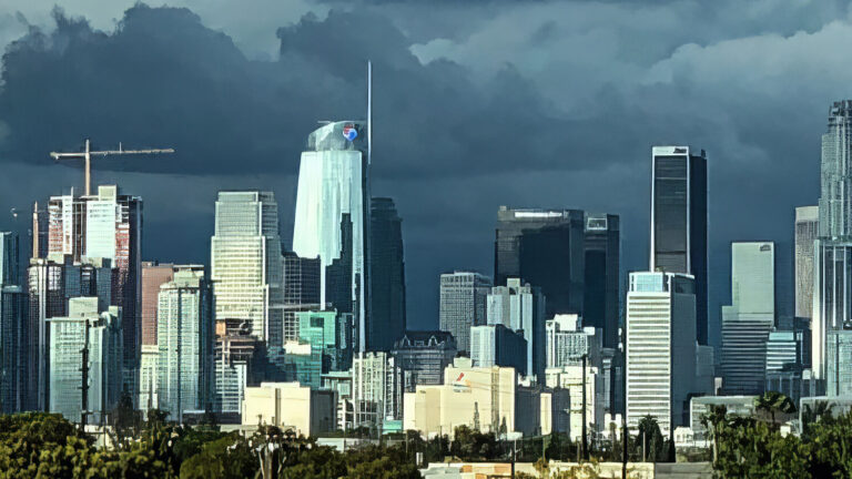 A panoramic view of downtown Los Angeles, featuring tall skyscrapers under a dramatic, cloudy sky, with palm trees and greenery visible in the foreground.