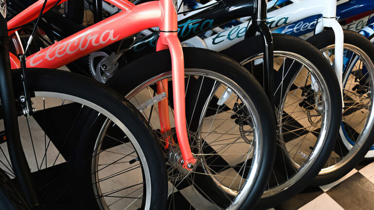 A row of Electra bicycles with wide tires is lined up on a black-and-white checkered floor, showing different frame colors and styles closely arranged side by side.