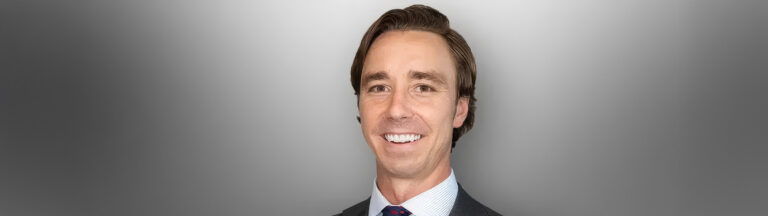 A smiling man with brown hair in a suit, shirt, and tie poses against a smooth gray gradient background.