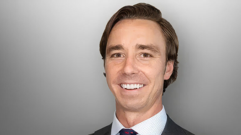 A smiling man with brown hair in a suit, shirt, and tie poses against a smooth gray gradient background.