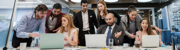 A group of business professionals gather around a table, collaborating and working on laptops in a modern office setting, with papers, pens, and drinks on the table.