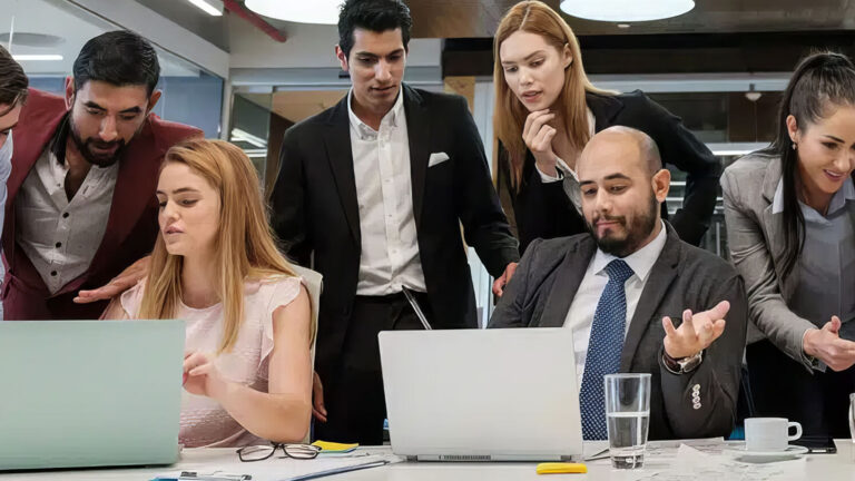 A group of business professionals gather around a table, collaborating and working on laptops in a modern office setting, with papers, pens, and drinks on the table.