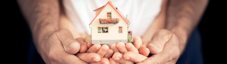 Close-up of adult hands gently holding a child’s hands, which are cupping a small model house. The scene symbolizes protection, care, and the concept of home and family.