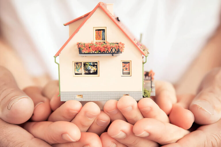 Close-up of adult hands gently holding a child’s hands, which are cupping a small model house. The scene symbolizes protection, care, and the concept of home and family.