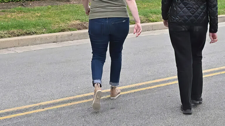 Two people walking side by side along the middle of a street, captured from behind. One wears jeans and sneakers, the other wears black pants and a black jacket. Grass and a curb are visible in the background.