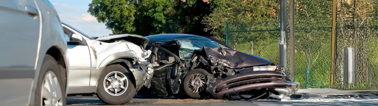 Two badly damaged cars after a collision, with crumpled front ends and debris on the road, next to a fence and trees in the background.