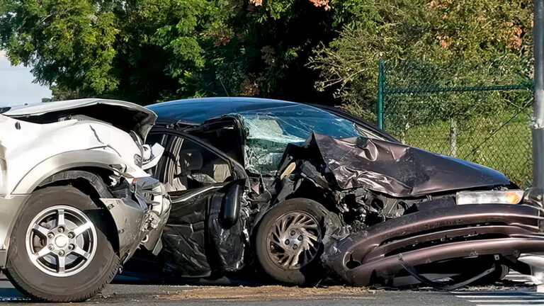 Two badly damaged cars after a collision, with crumpled front ends and debris on the road, next to a fence and trees in the background.