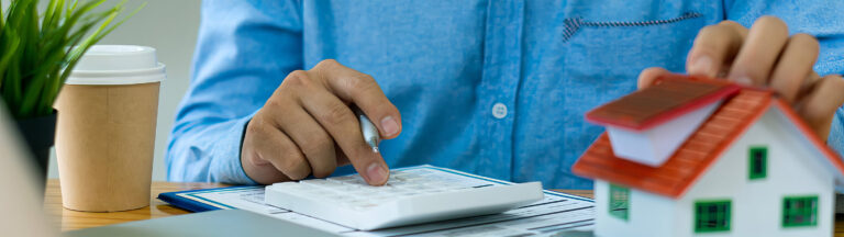 A person in a blue shirt uses a calculator next to a small model house, paperwork, and a coffee cup, suggesting financial calculations related to real estate or home buying.