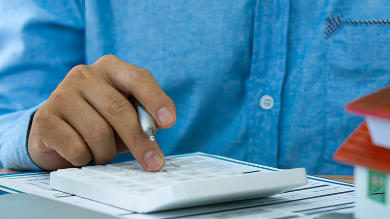 A person in a blue shirt uses a calculator next to a small model house, paperwork, and a coffee cup, suggesting financial calculations related to real estate or home buying.