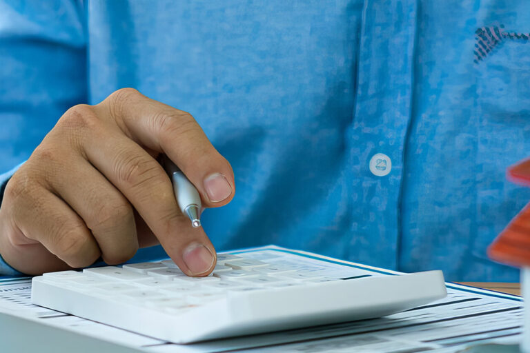 A person in a blue shirt uses a calculator next to a small model house, paperwork, and a coffee cup, suggesting financial calculations related to real estate or home buying.