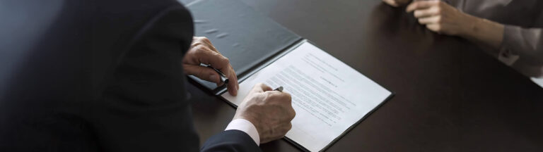 A person in a suit signs a document on a clipboard at a dark desk, while another person sits across from them with hands folded.