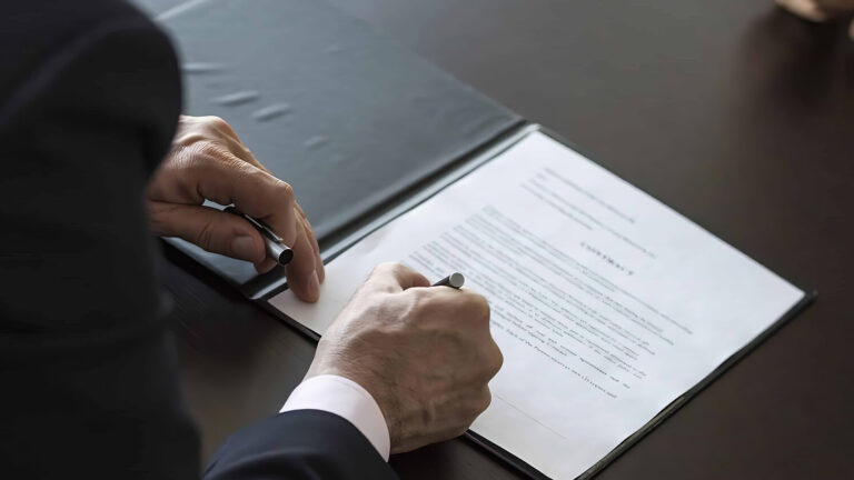 A person in a suit signs a document on a clipboard at a dark desk, while another person sits across from them with hands folded.