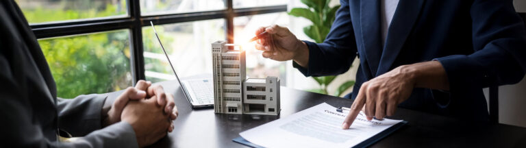 Two people in business attire sit at a desk with a laptop, miniature building models, and a contract. One person gestures with a pen while the other points to the document.