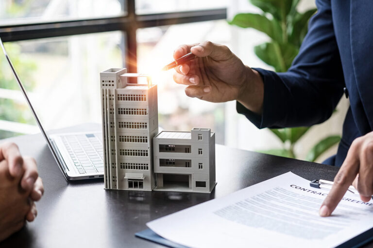 Two people in business attire sit at a desk with a laptop, miniature building models, and a contract. One person gestures with a pen while the other points to the document.